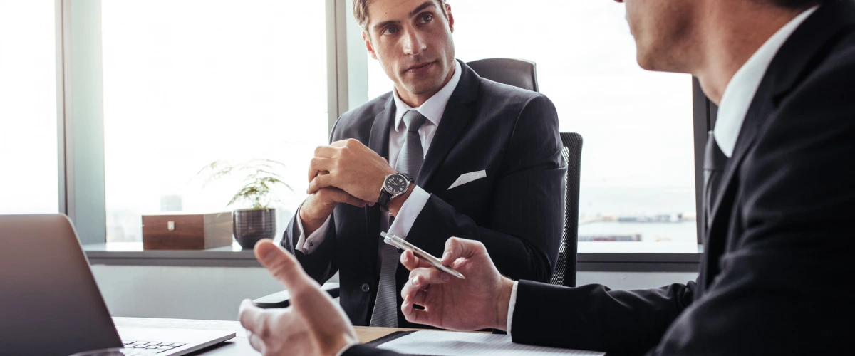 Two businessmen in suits discussing in an office, one holding a pen and listening intently, the other gesturing