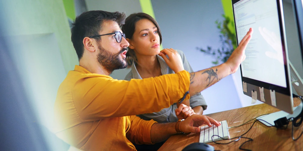 A man with a beard and glasses pointing at a computer monitor while a woman looks on, both engaged with code or data.