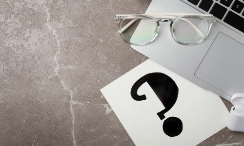 A piece of paper with a large black question mark on a marbled desk next to a laptop, clear glasses, and wireless earbuds.