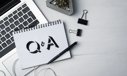 A spiral notebook with "Q & A" written on it, a black pen, paper clips, a small succulent plant, and a laptop on a white desk.