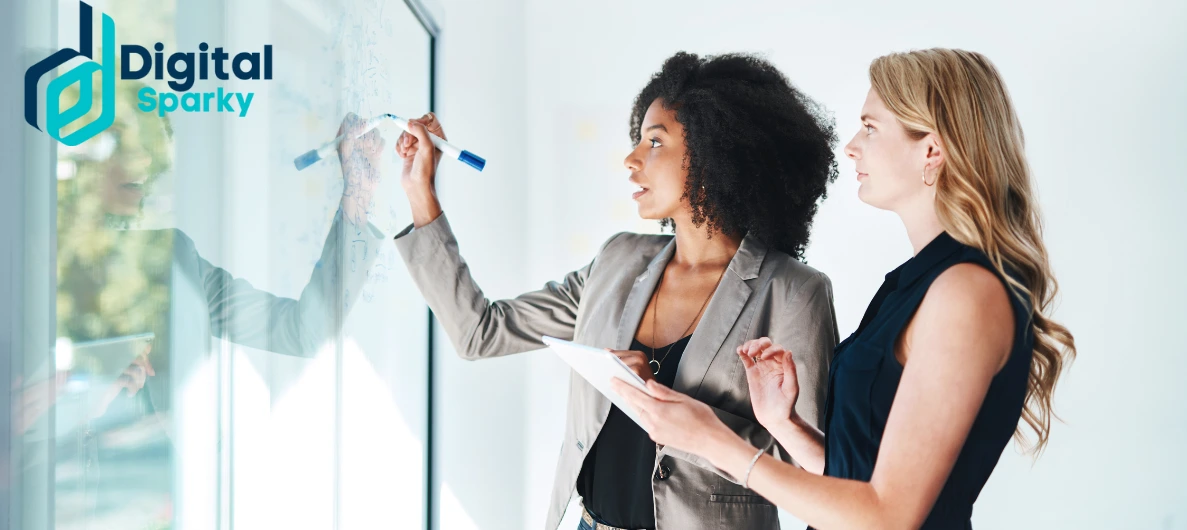 Two professional women collaborating and writing on a transparent whiteboard, with the Digital Sparky logo in the corner.