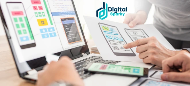 Two people collaborating on mobile app design and development, reviewing wireframes on paper and code on a laptop, with the Digital Sparky logo.