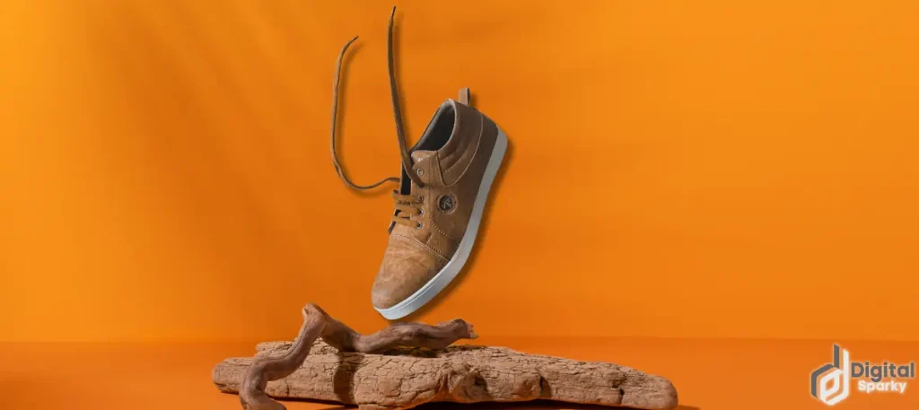 A single brown leather casual shoe with its laces untied, suspended above two pieces of driftwood against a vibrant orange background, with the Digital Sparky logo.